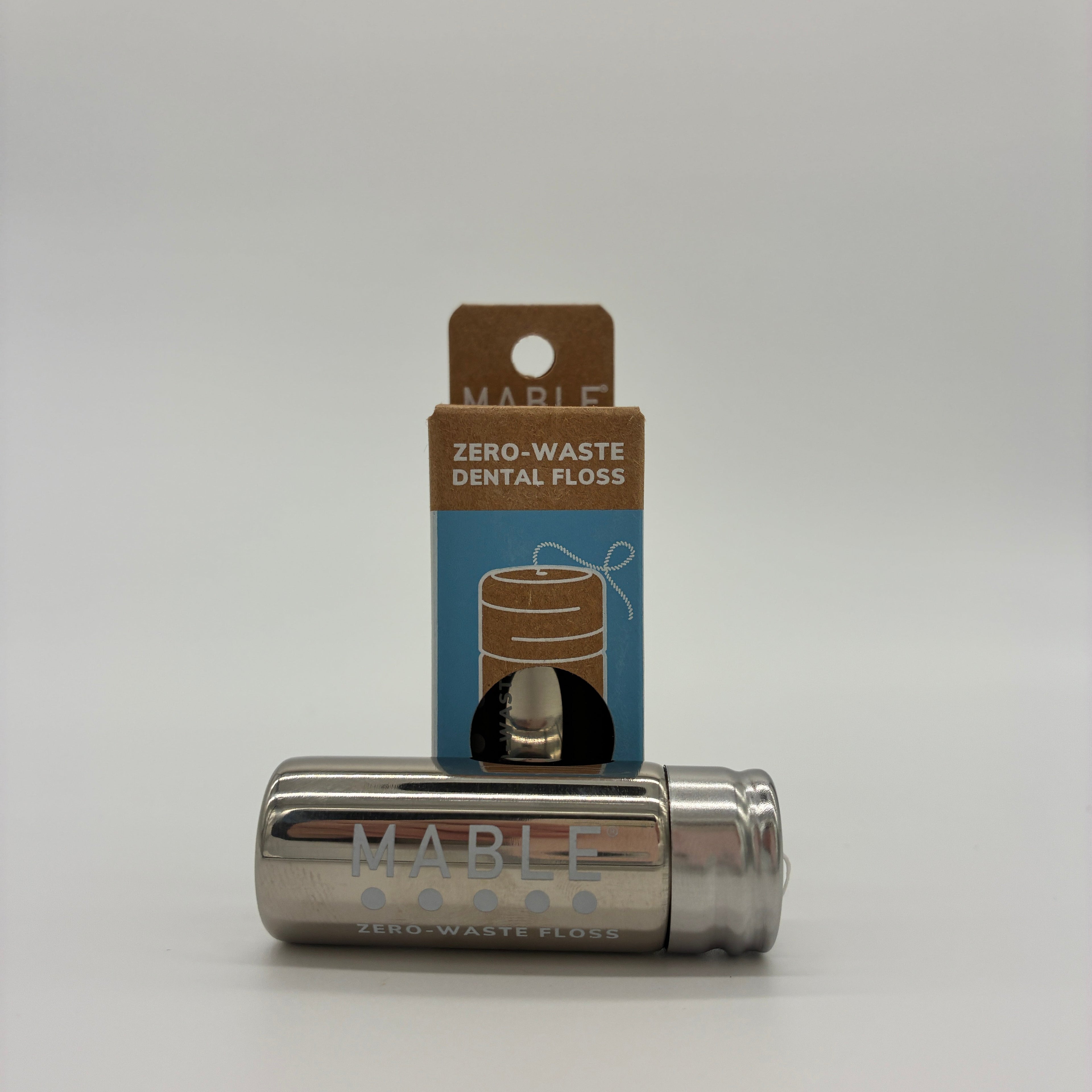 A metallic silver Mable Zero-Waste Dental Floss tube lying horizontally in front of its blue and brown cardboard packaging card, set against a seamless light gray background.