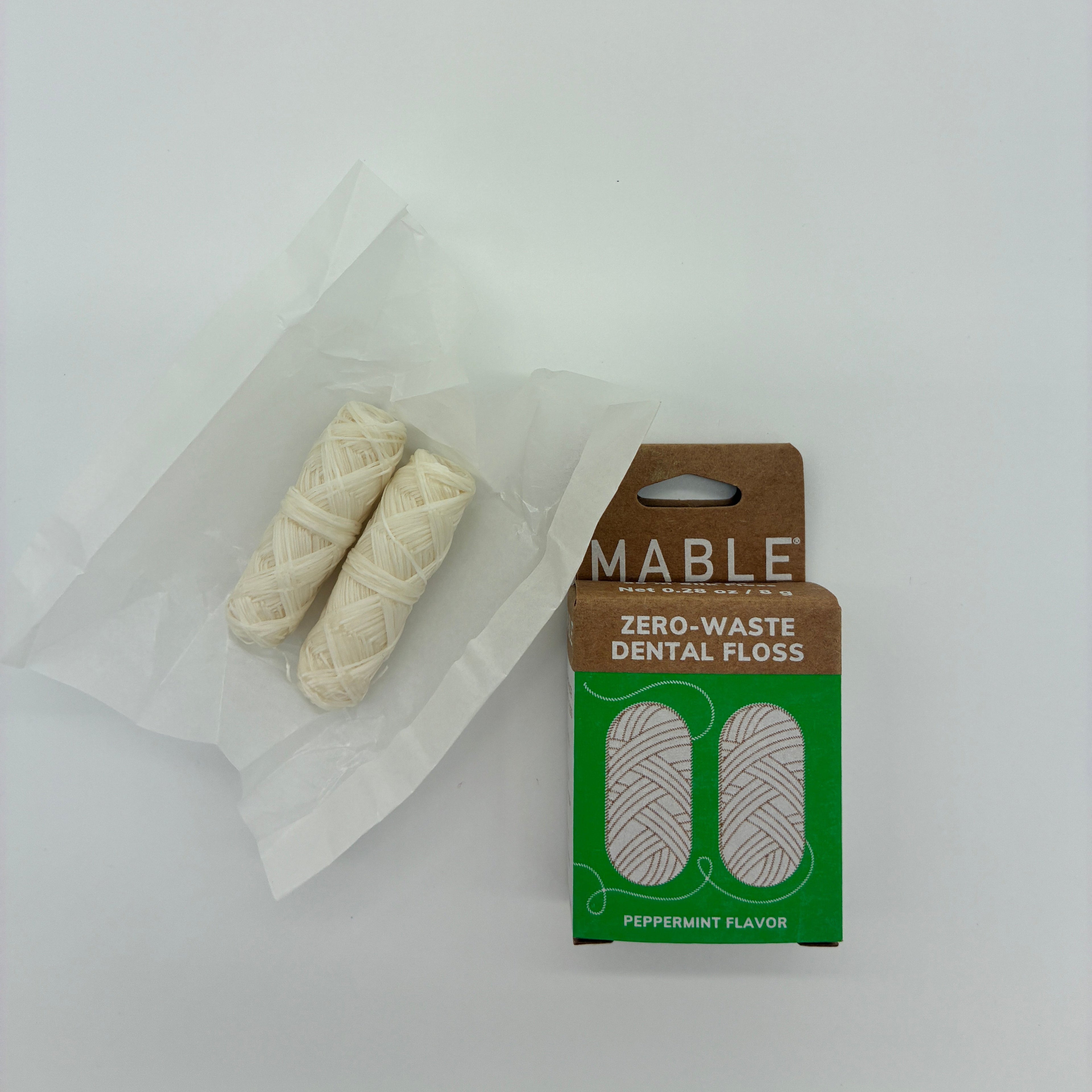 Two off-white dental floss rolls in a clear plastic bag next to their brown and green cardboard packaging box labeled 'MABLE Zero-Waste Dental Floss Peppermint Flavor'.