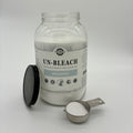 A clear glass bottle of oxygen powder with its black lid placed beside it and a metal scoop filled with the white powder on a light gray background.